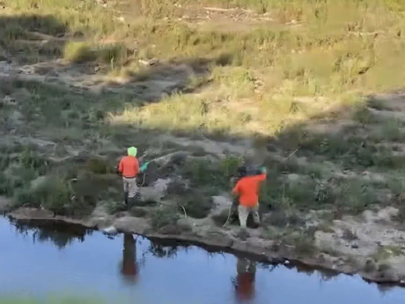 Two workers in orange tops on a creek bank above shallow water, one of them spraying vegetation with a handheld wand.